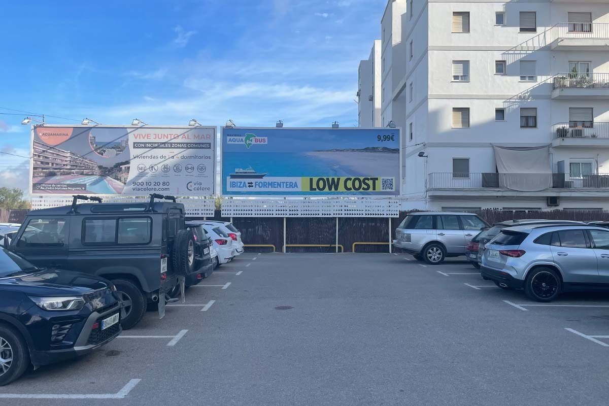 Aparcamiento junto al puerto de Ibiza con coches y, detrás, dos vallas publicitarias contra un edificio; en la de la derecha, Naviera Aquabus anuncia la travesía Ibiza – Formentera como low cost por 9,99 euros, con un código QR al lado