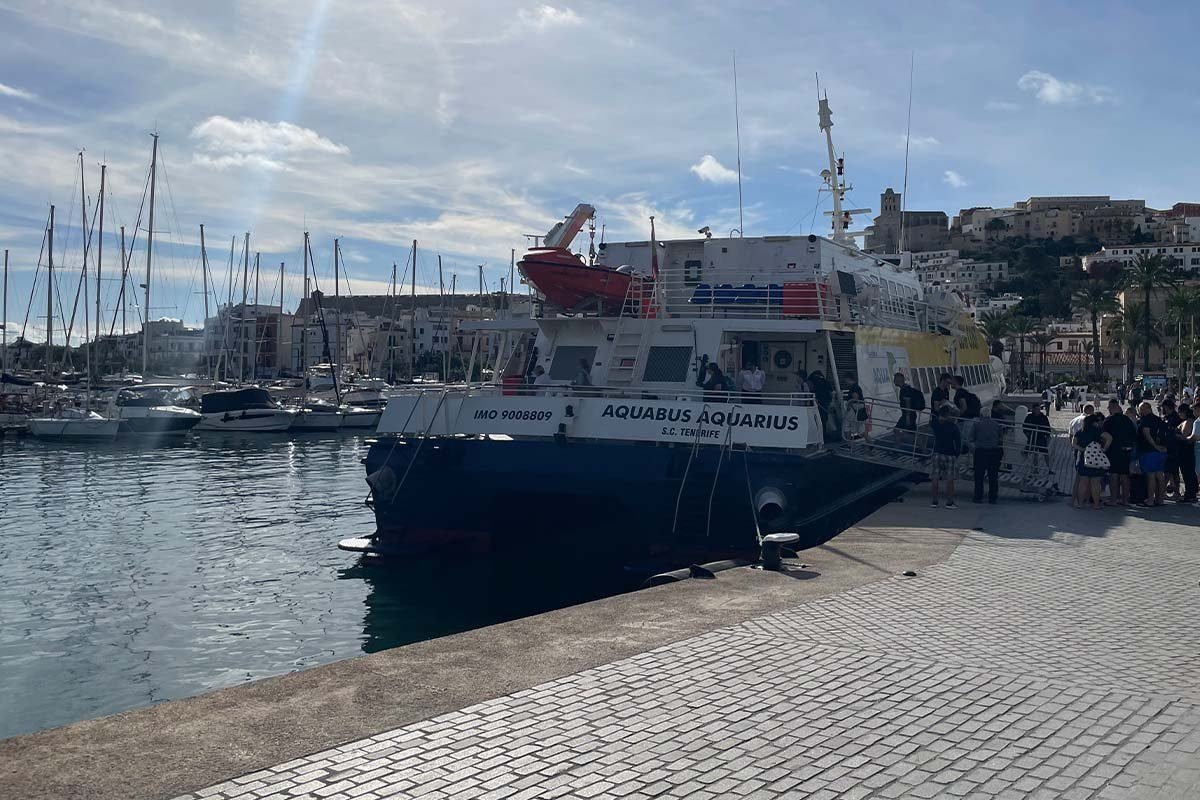 El ferry rápido Aquabus Aquarius, de casco azul oscuro, en el muelle del puerto de Ibiza, con una fila de pasajeros esperando al lado y la ciudad alta de Dalt Vila en la colina detrás