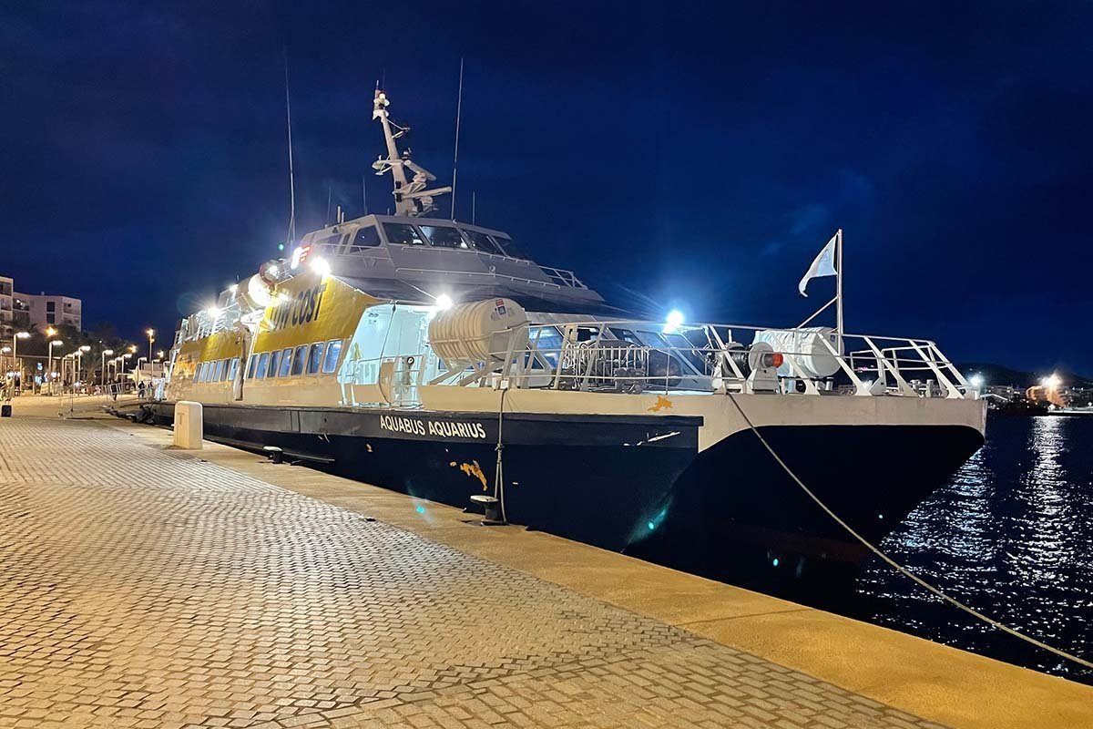 El ferry Aquabus Aquarius, con la superestructura amarilla, amarrado de noche en un muelle iluminado, con las luces de cubierta encendidas bajo un cielo azul oscuro