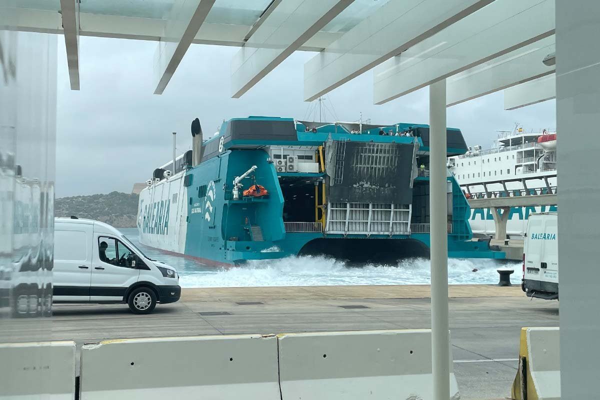 Un ferry de Baleària de casco blanco y popa turquesa maniobra junto al muelle levantando agua blanca, con la rampa de popa ya abierta y un bote salvavidas naranja, visto desde una zona de espera cubierta con una furgoneta de Baleària en el muelle y otro barco blanco detrás