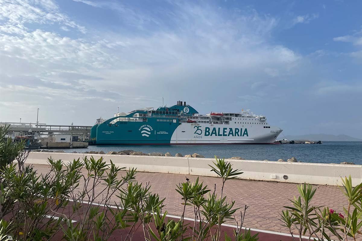 El ferry Bahama Mama de Baleària junto a un muelle bajo un cielo azul, con la proa turquesa, el casco blanco con el rótulo 25 años Baleària y el distintivo verde Rumbo Verde con el texto impulsado por gas natural, visto desde un paseo con adelfas en primer plano