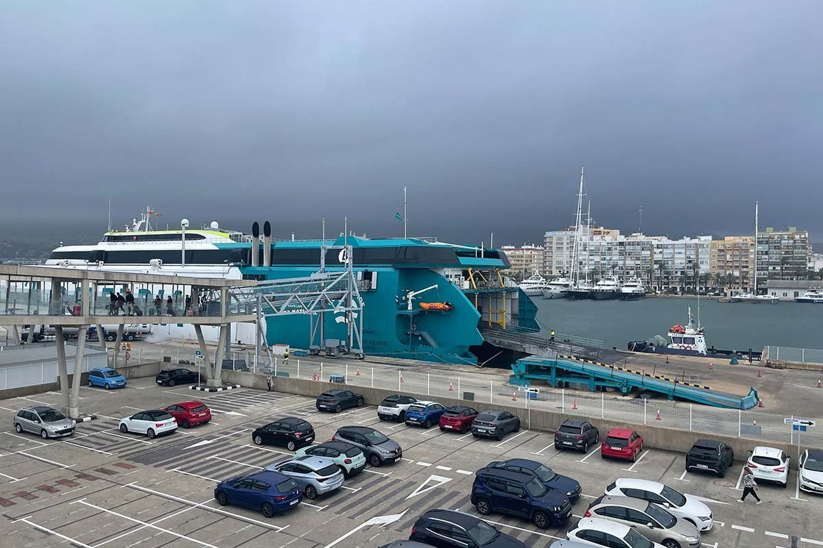 Vista sobre el aparcamiento lleno del puerto de Dénia hacia un ferry rápido turquesa de Baleària con la rampa de popa abierta, una pasarela cubierta por la que pasan pasajeros con maletas y, detrás, el puerto deportivo y los edificios del pueblo bajo un cielo gris