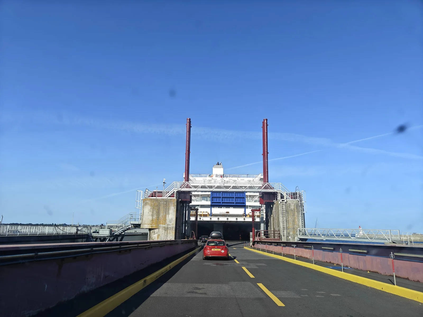 Un coche rojo sube por la rampa a la proa abierta de un ferry blanco de Brittany Ferries, bajo un cielo azul, en el puerto de Saint-Malo