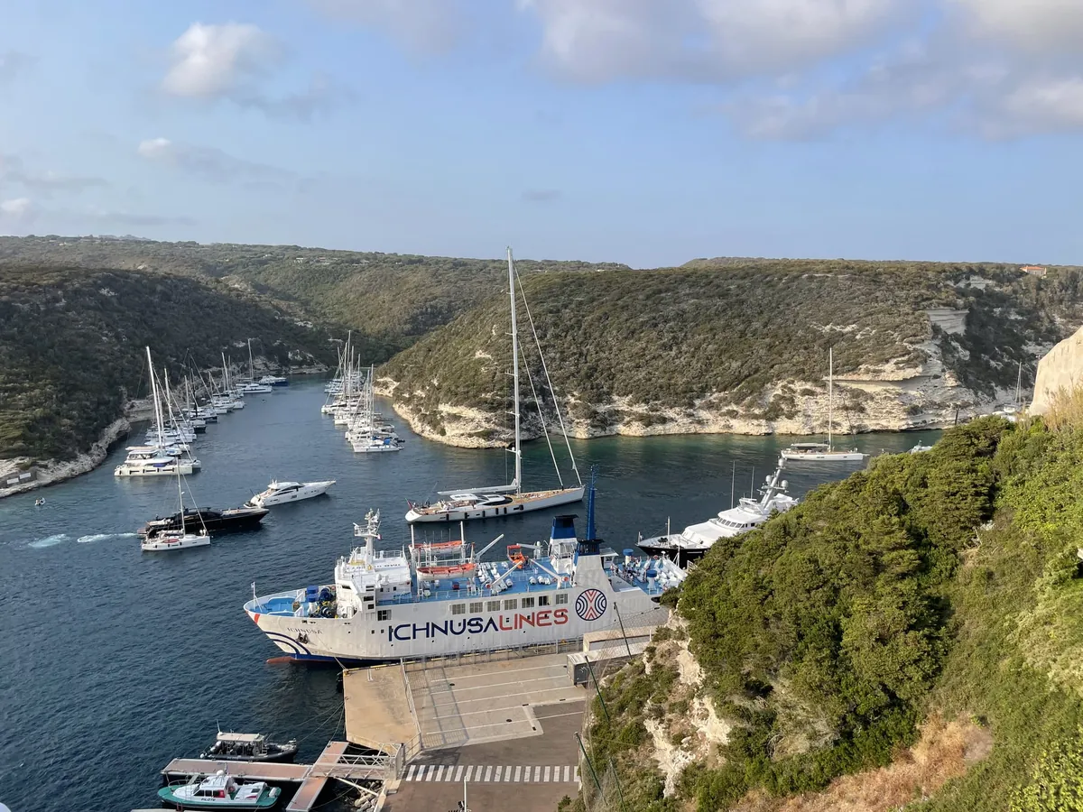 El ferry blanco Ichnusa de Ichnusa Lines atracado en el muelle de Bonifacio, visto desde arriba, en una ensenada estrecha entre acantilados blancos de caliza con vegetación, con decenas de veleros y lanchas en el agua