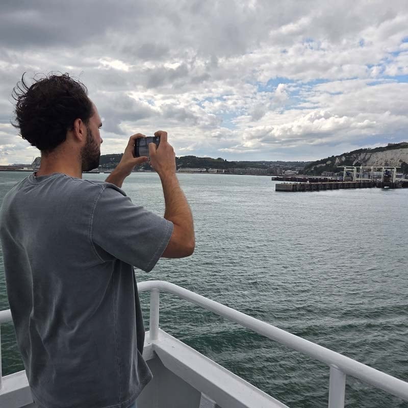 Bram Tigchelaar con camiseta gris hace una foto con el móvil desde la barandilla de un ferry, delante de él el puerto y los blancos acantilados de tiza de Dover