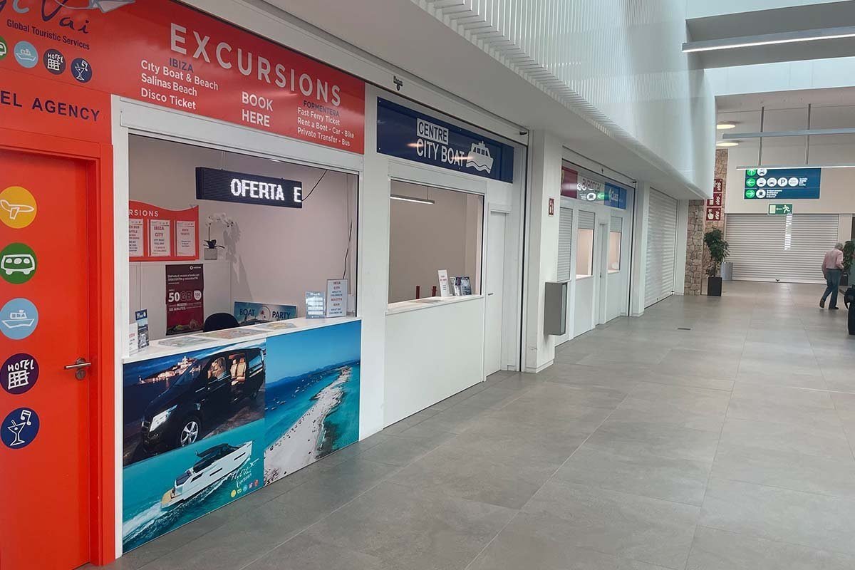 Fila de taquillas en el vestíbulo de la terminal de ferris de Ibiza: delante, una agencia roja que ofrece excursiones, billetes de barco rápido a Formentera, alquiler de coches y bicis y traslados; al lado, una taquilla azul de Centre City Boat y, más allá, persianas cerradas