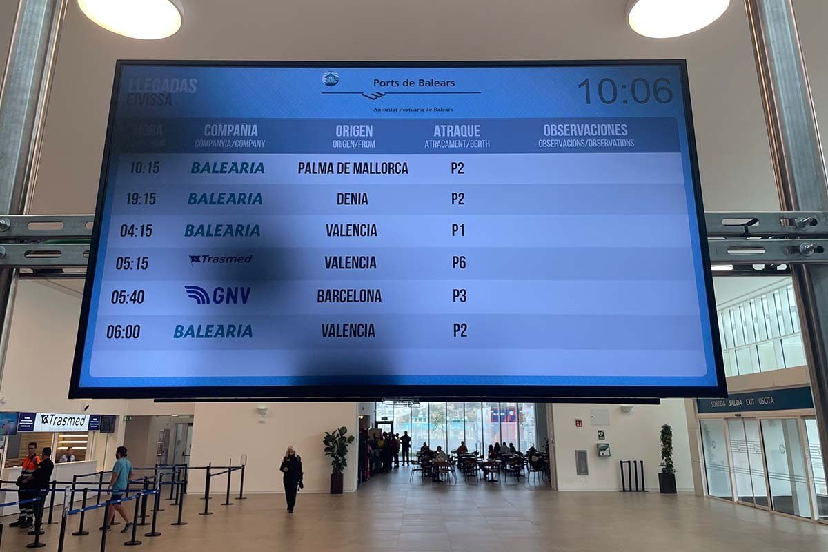 Gran panel de llegadas de Ports de Balears en la terminal de ferris de Ibiza, con el título LLEGADAS EIVISSA y seis llegadas: Baleària desde Palma de Mallorca, desde Dénia y dos veces desde Valencia, Trasmed desde Valencia y GNV desde Barcelona, cada una con su atraque, de P1 a P6; debajo, un mostrador de Trasmed y la salida
