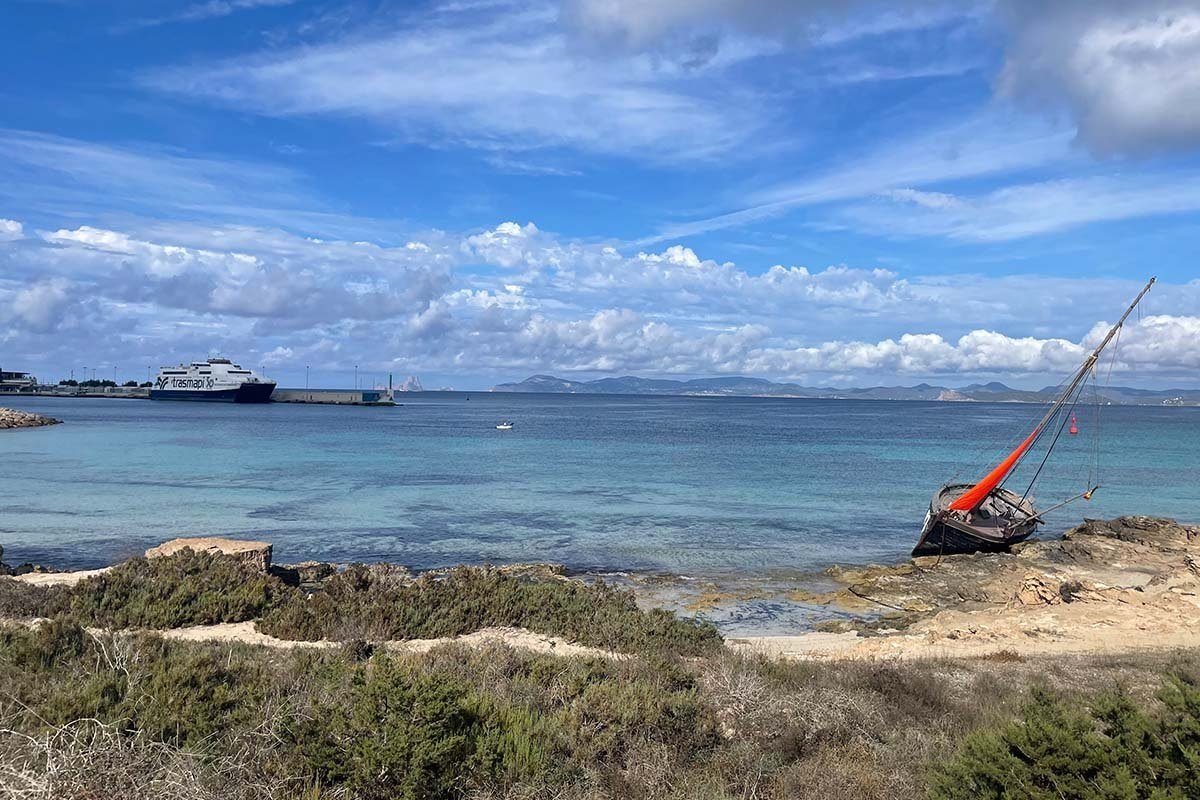 Vista desde la costa rocosa de Formentera sobre un agua turquesa y transparente hacia un ferry de Trasmapi junto al dique de La Savina, con la costa de Ibiza en el horizonte y, a la derecha, una barca de pesca varada en las rocas