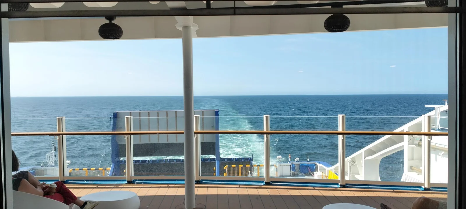 Vista desde la cubierta de popa cubierta del ferry, por encima de la barandilla, sobre la estela y el mar, con un pasajero en un sillón blanco a la izquierda