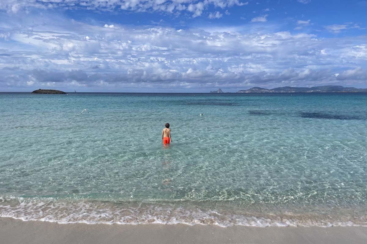 Un niño con bañador naranja de pie en el agua poco profunda y transparente, junto a una playa de arena blanca, con un islote en el horizonte y la costa de Ibiza al fondo