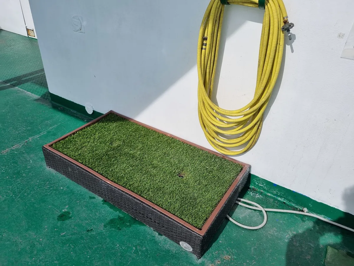 Una bandeja rectangular de césped artificial con marco negro sobre una cubierta exterior verde, con una manguera amarilla enrollada en la pared blanca de encima