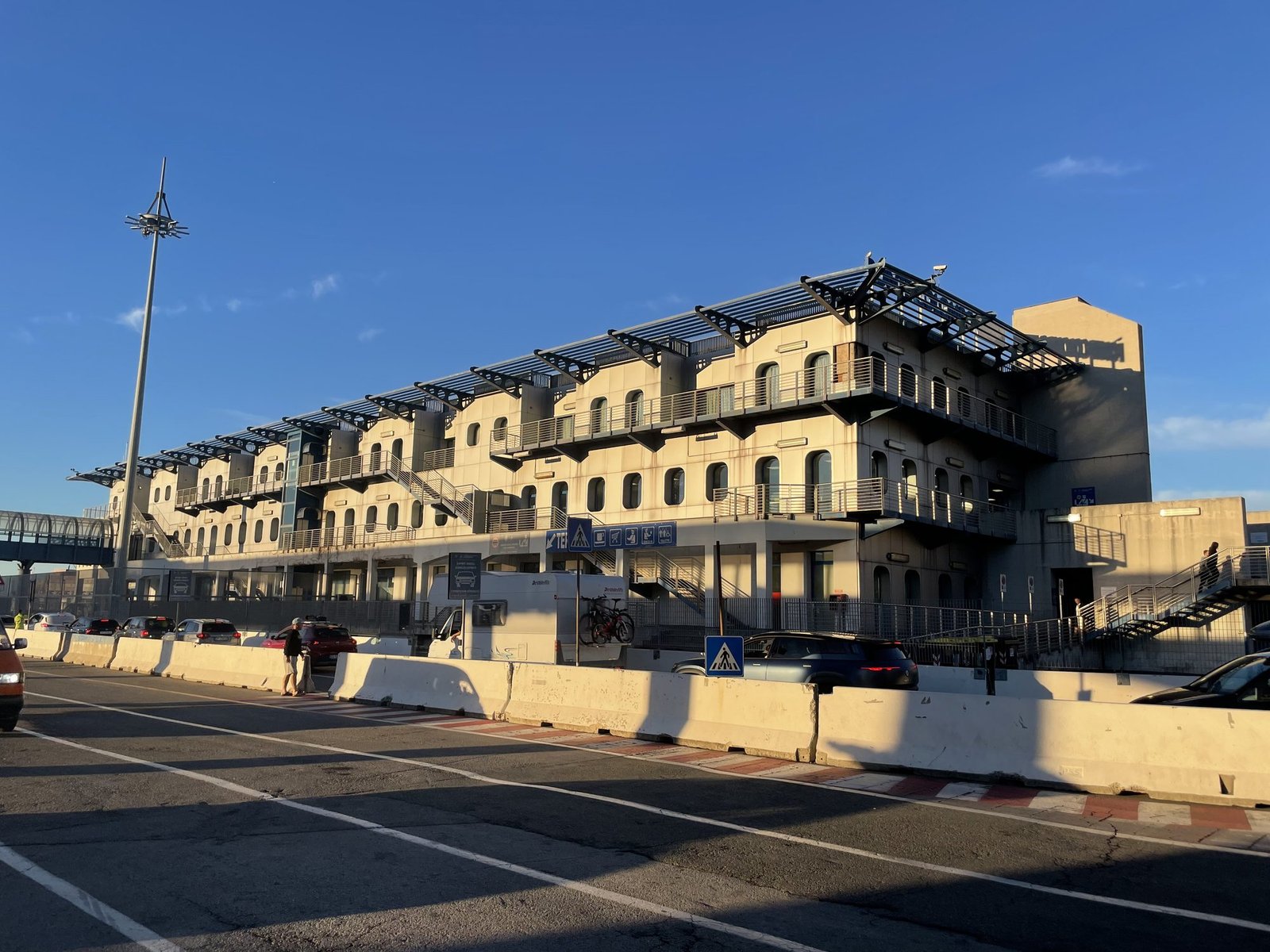 El edificio de la terminal de ferris del puerto de Génova al sol de primera hora, con una fila de coches y una autocaravana con bicicletas delante