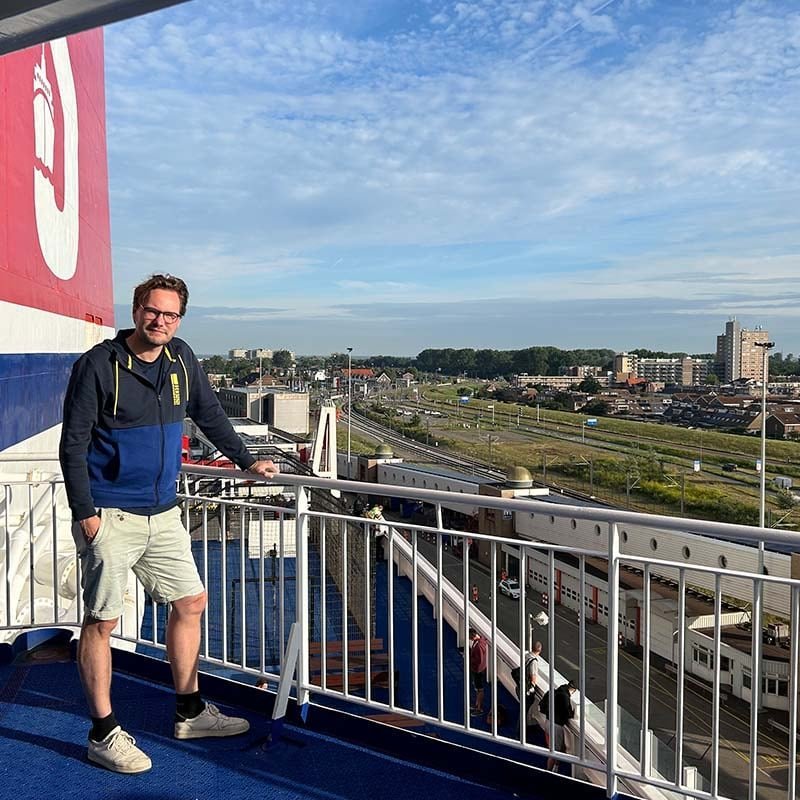 Jelle van der Bij, con gafas, una sudadera azul con capucha y pantalón corto, en la cubierta exterior de un ferry, apoyado en la barandilla, a la izquierda la chimenea roja y abajo una ciudad portuaria con vías de tren