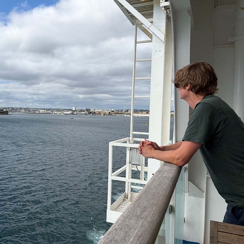 Jochem Vroom con camiseta verde, apoyado en la barandilla de madera de un ferry, mira el agua y una ciudad costera bajo un cielo nublado