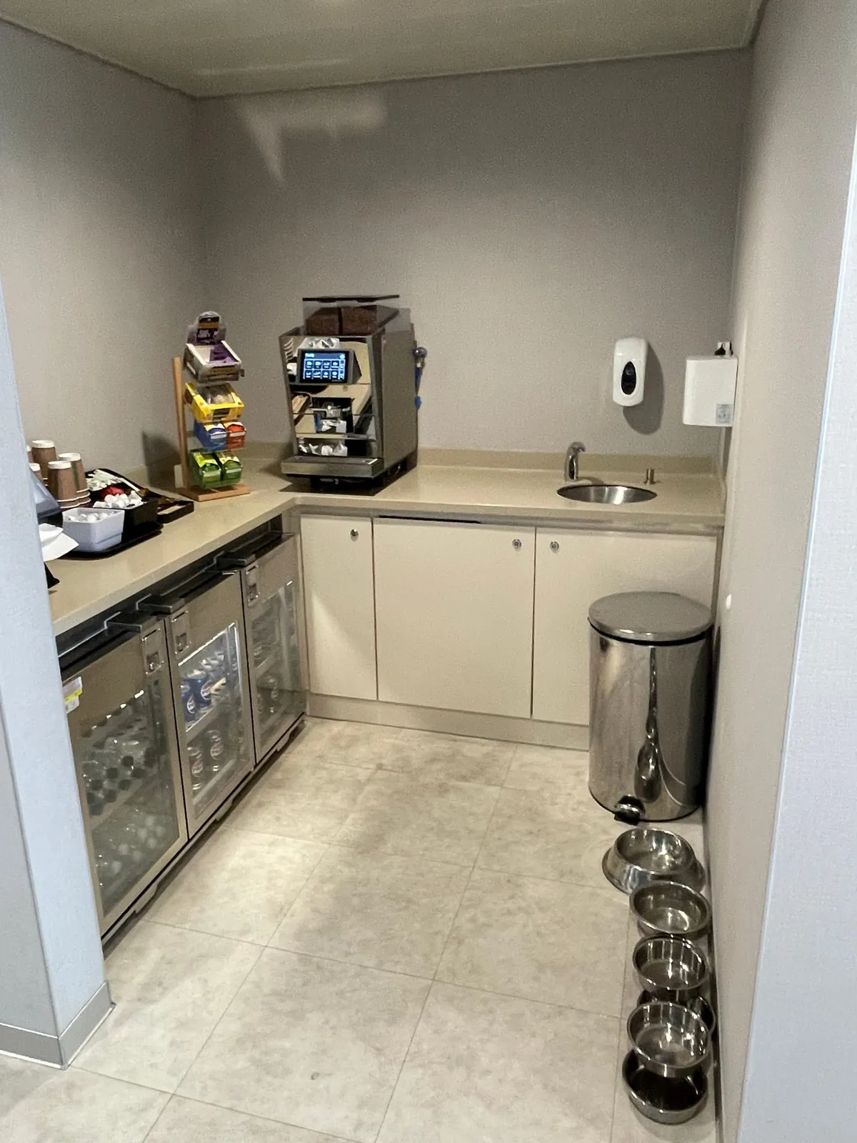 Un rincón de cocina con una cafetera, neveras con latas y botellas, vasos y golosinas sobre la encimera, un fregadero, una papelera y en el suelo una fila de cuencos de acero inoxidable