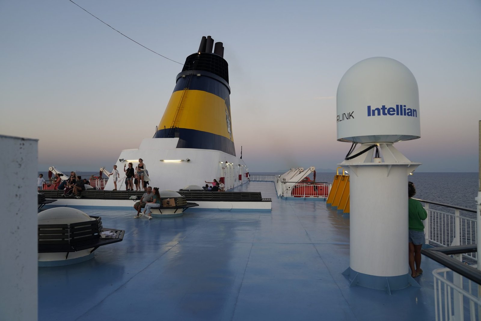 La cubierta superior del ferry al atardecer, con la chimenea amarilla, pasajeros en bancos y una cúpula de satélite a la derecha